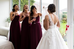 three bridesmaids smiling at a bride in her wedding dress. The brdiesmaids are wearing maroon dresses and they are in front of a window.
