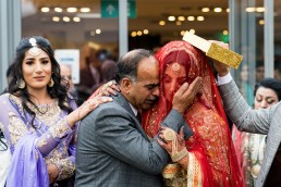 asian bride in a red veil comforted by her father as she leaves her wedding