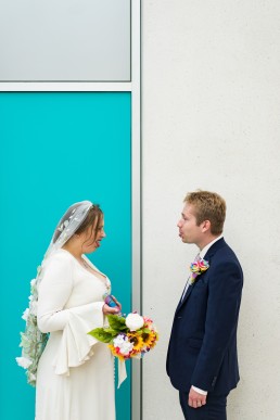 bride and groom pull faces at each other in front of a teal wall panel at Millenium gallery in Sheffield