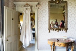 wedding dress hangs in a doorway as bridesmaids get ready in the mirror reflection
